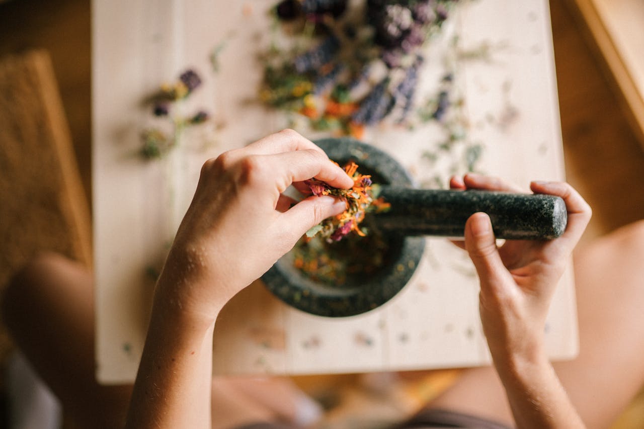 A close-up view of hands grinding herbs and flowers in a mortar and pestle on a wooden table.